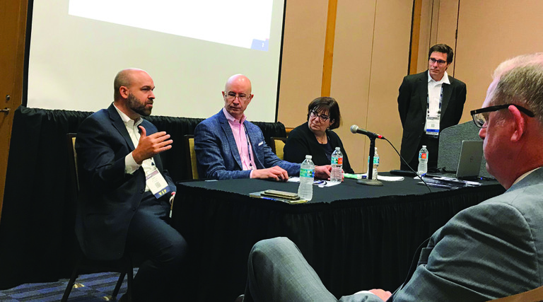 Two bald men sitting at a table next to a woman, one man standing at a podium, one man sitting and listening