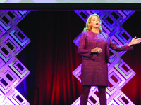 blond woman in cranberry colored suit gesturing on a stage