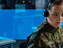 Woman with headset on, in a room with other people working on computers.